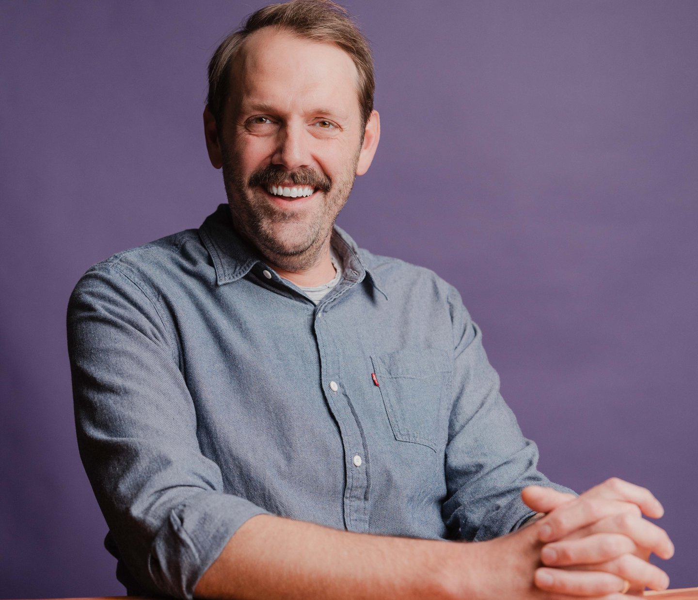 Richard Whitfield, Senior Software Engineer at Dozen Software, smiling with arms folded at a wooden table against a purple backdrop
