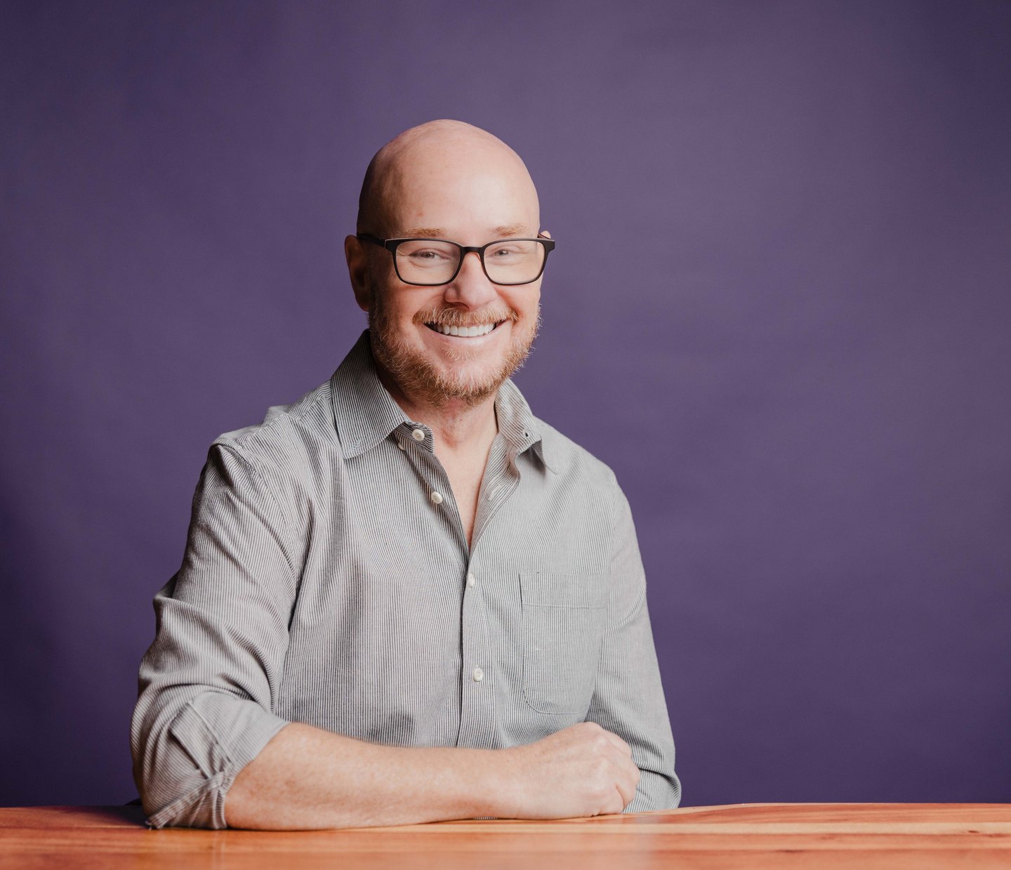 Chris Downs, Founder and CEO of Dozen Software, smiling in glasses at a wooden table against a purple backdrop