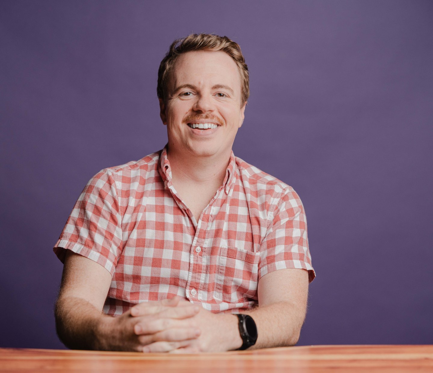 Jacob Lewis, Senior Software Engineer at Dozen Software, smiling in a checkered shirt at a wooden table against a purple backdrop