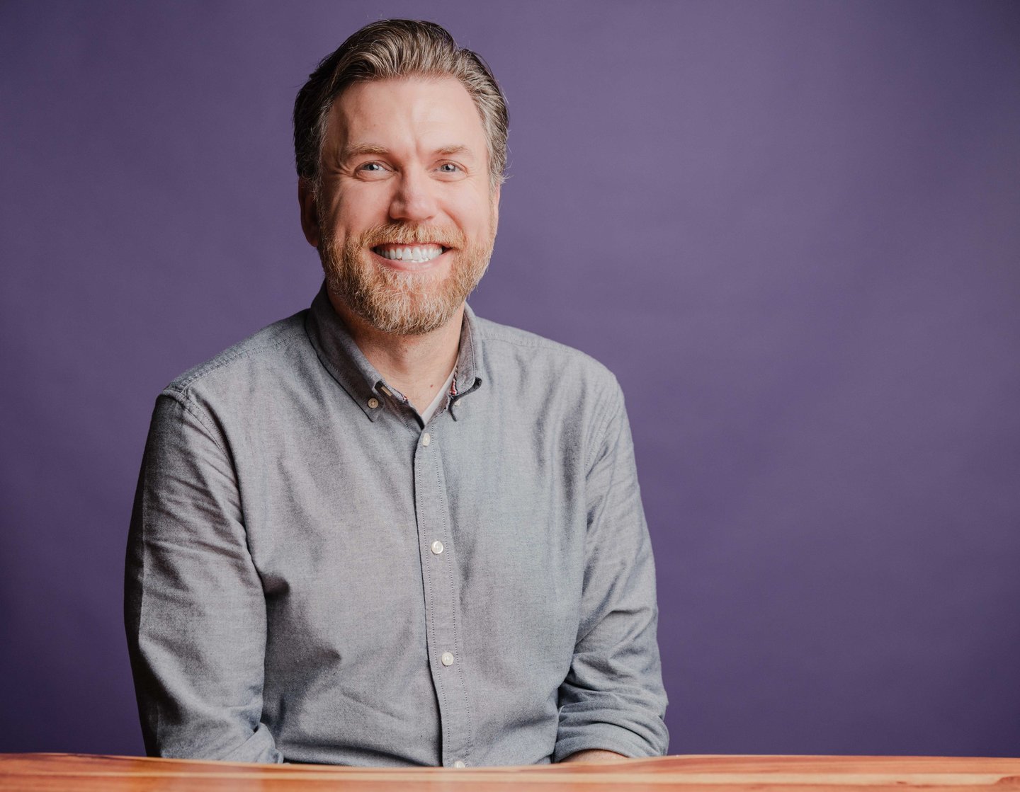 Chris Yommer, Principal Systems Engineer at Dozen Software, smiling with a gray beard at a wooden table against a purple backdrop