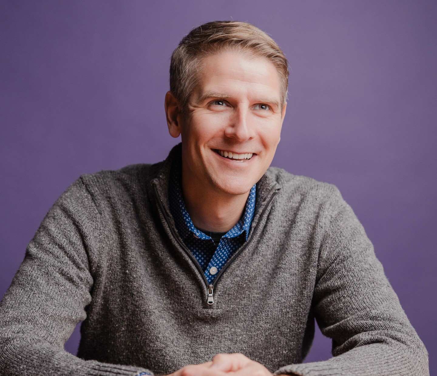 Clay Wittwer, Managing Partner of Engineering and Operations at Dozen Software, smiling in a gray sweater at a wooden table against a purple backdrop