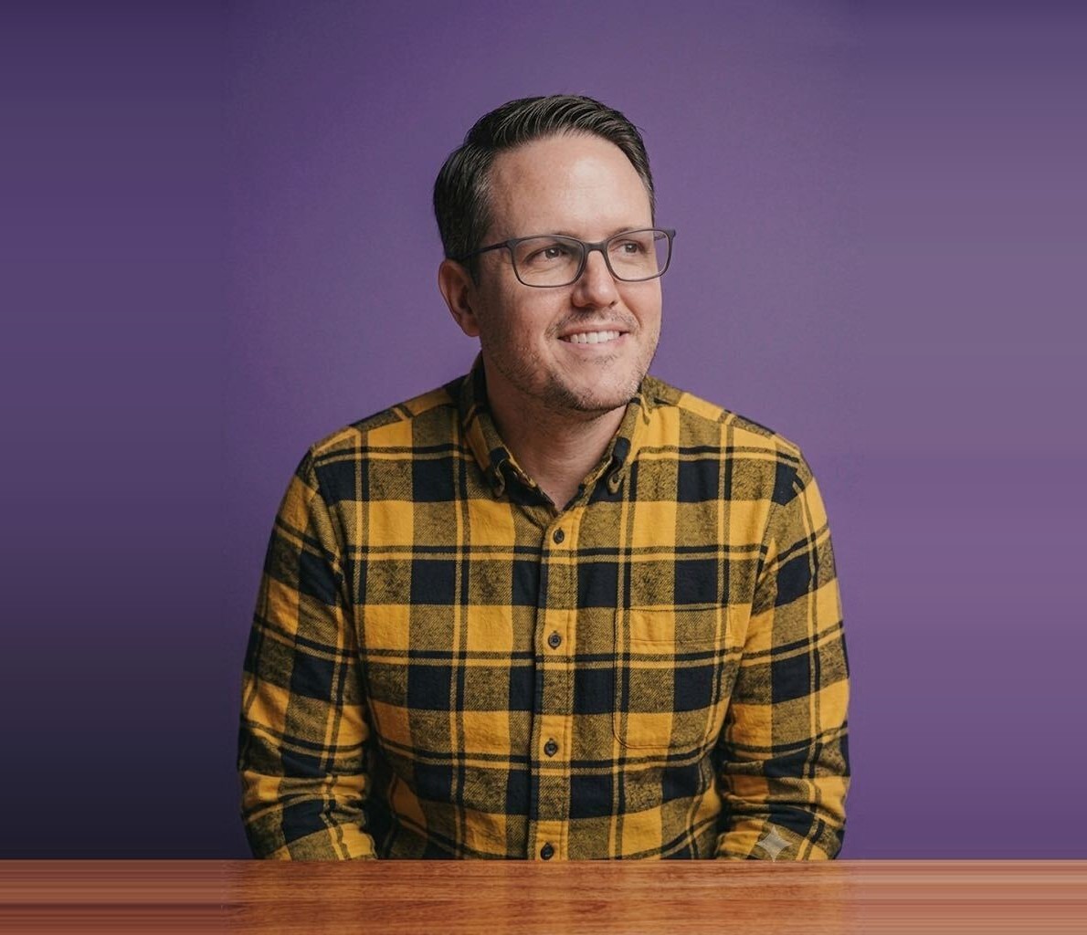 Chris Reising, Principal Software Engineer at Dozen Software, smiling in glasses and a plaid shirt at a wooden table against a purple backdrop